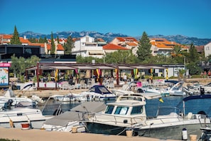 A marina with boats docked, a covered seating area, and buildings with red roofs in the background.