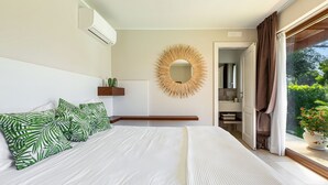A bedroom with a large bed, a round mirror, a small shelf with plants, and a view of greenery through the window. - 3680 Villa Monica by Perle di Puglia (Polignano a Mare)