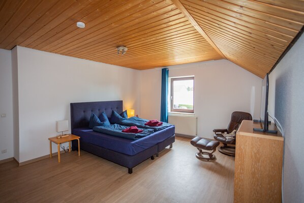 A bedroom with a wooden ceiling, a bed with blue bedding, a small table, a chair, and a desk.