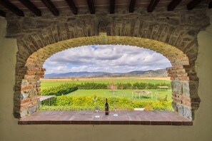 A stone archway leading to a terrace with a bottle and glasses, overlooking a green landscape with a clear sky.