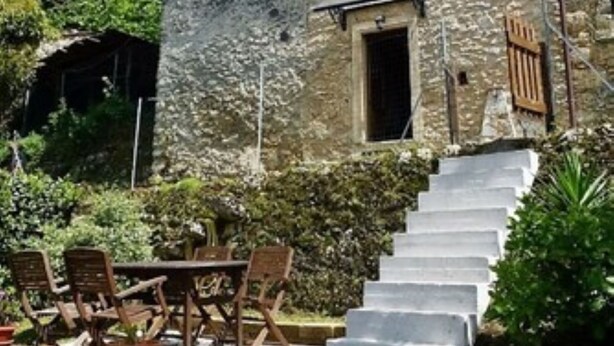 Un bâtiment en pierre avec un escalier menant à une porte, entouré de verdure et de mobilier d’extérieur.