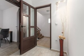 A room with a wooden door, a chair, a vase with branches, and a staircase in the background.