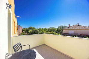 A balcony with a table and chairs, a clear blue sky, and a view of neighboring buildings and greenery.