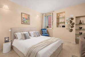 Un dormitorio con cama, mesita de noche, silla, ventana y estanterías en la pared.