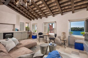 A cozy living area with a sofa, a dining table with chairs, a kitchen with a built-in oven, and a view of the sea through a window. - Natascha’s Beachfront/Stylish Two-Story Apartment A2 (Rovinj)