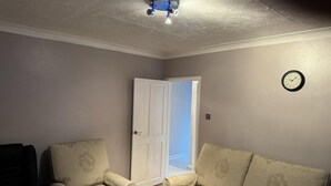Une pièce avec deux fauteuils beiges, une porte blanche, une horloge murale et un luminaire au plafond.