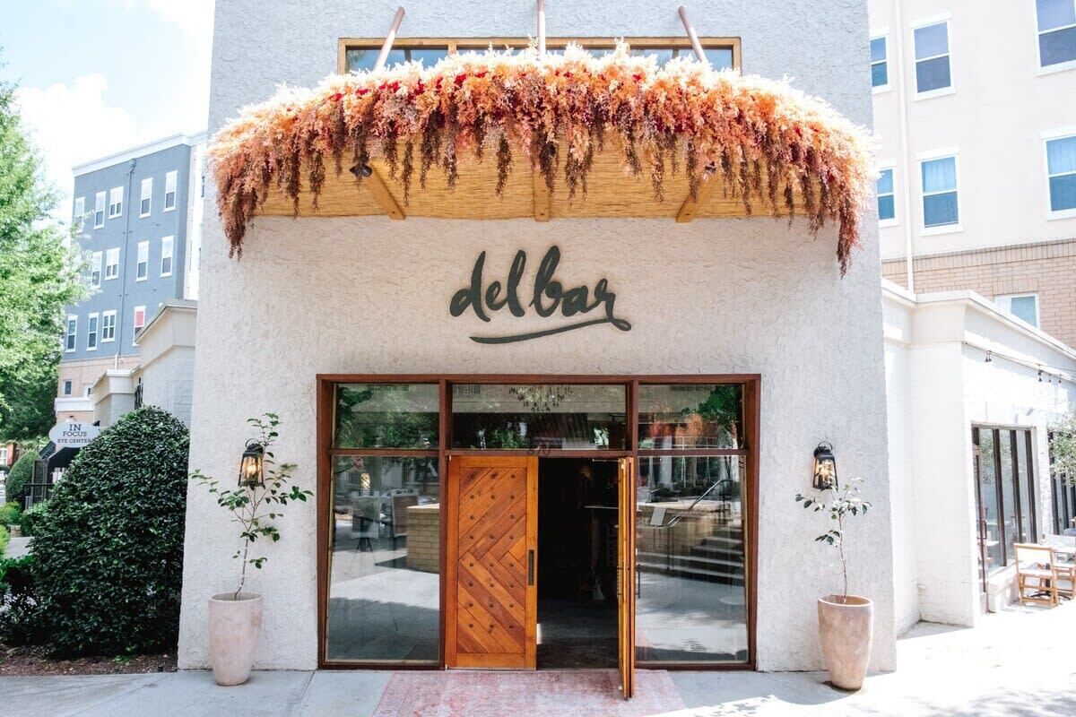 Un bâtiment avec une porte en bois et le mot « delbar » écrit au-dessus. De chaque côté de l’entrée, on peut voir des plantes en pot.