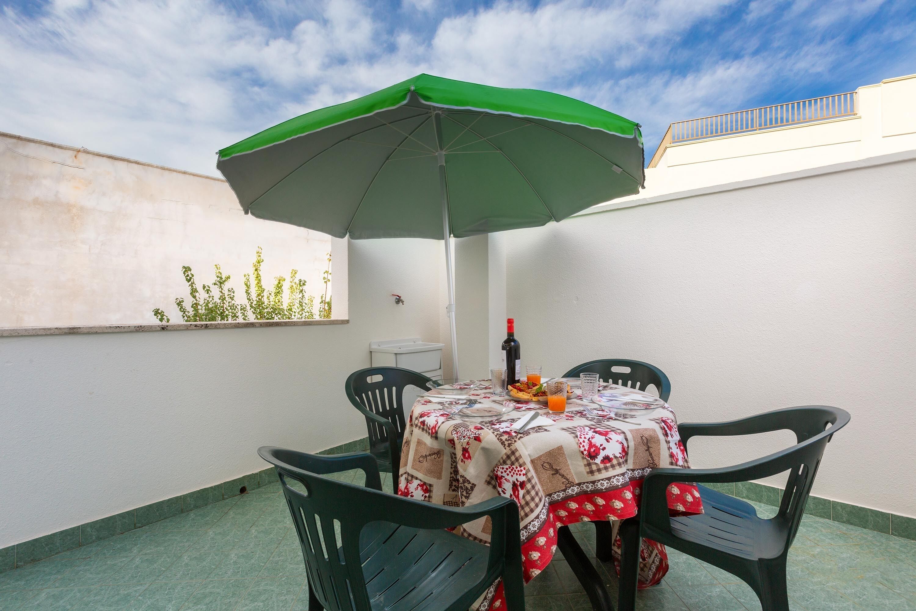 A small outdoor patio with a table and chairs, a green umbrella, and a bottle of wine.