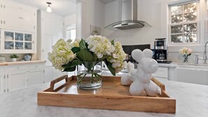 Una cocina con armarios blancos, una campana extractora de acero inoxidable y una bandeja de madera con un jarrón de flores y una escultura con globos.
