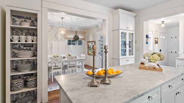 Una cocina moderna con armarios blancos, encimera de mármol y comedor con mesa y sillas blancas.