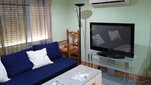 Een woonkamer met een blauwe bank, een televisie op een glazen standaard, een airconditioner en een vloerlamp.