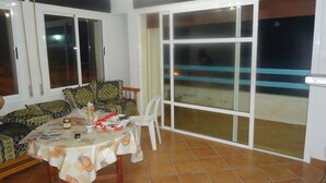 A dining area with a table covered by a tablecloth, a sofa, and a chair. There are windows and a sliding glass door leading to an outdoor area. - Martil apartment facing the sea (Martil)