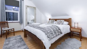 A bedroom with a wooden bed, a chair, a nightstand, a mirror, and a window with curtains. - Cosy Flat In London  (London)