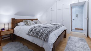 A bedroom with a wooden headboard, a large bed with white bedding, a grey blanket, and a bedside table with a lamp. There are white wardrobes and a bathroom visible through an open door. - Cosy Flat In London (London)