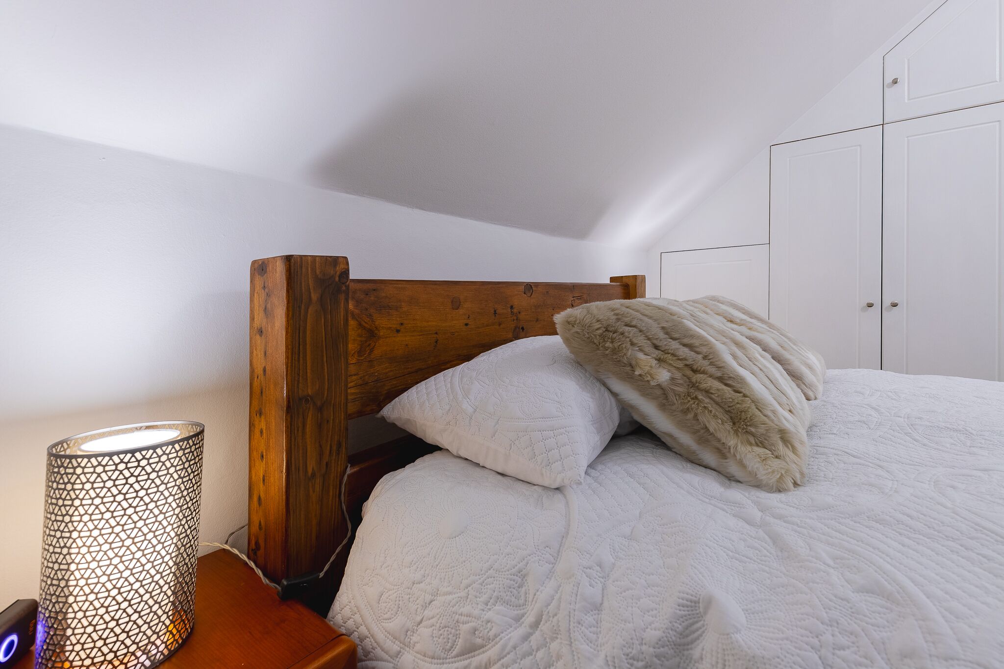 A bedroom with a wooden headboard, a white bed with a fluffy pillow, and a bedside lamp.