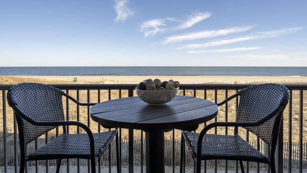 Une table ronde avec un saladier de fruits et deux chaises sur un balcon donnant sur une plage.