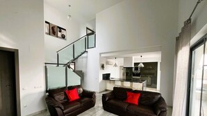 A modern living room with two leather sofas, a glass staircase, a kitchen in the background, and a large window.