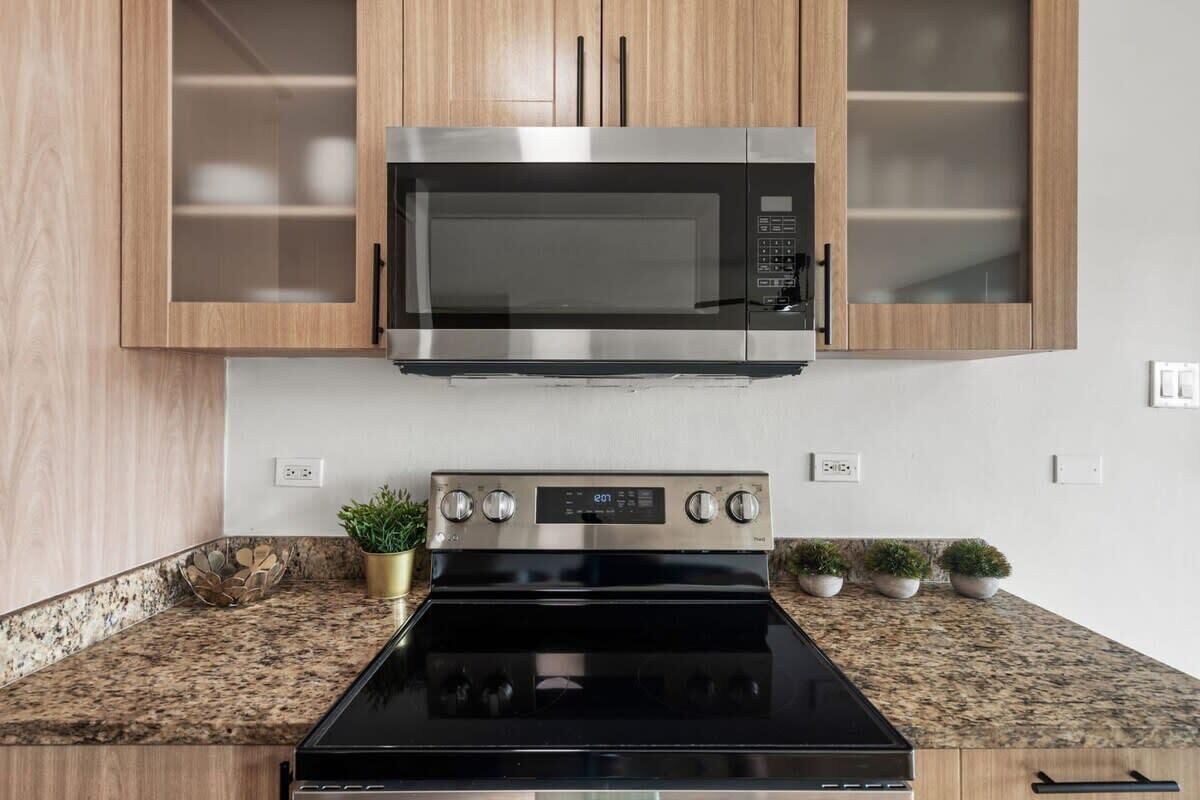 A modern kitchen with a stainless steel microwave and oven, wooden cabinets, granite countertop, and small potted plants.