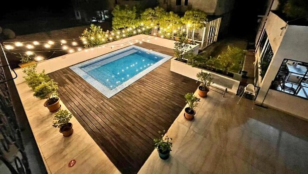 Une piscine extérieure entourée d’une terrasse en bois et agrémentée de plantes en pot, le tout mis en valeur par un décor de bâtiments et de guirlandes lumineuses.
