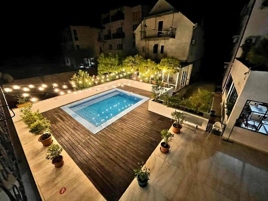 Piscina esterna circondata da un solarium in legno e piante in vaso, con sullo sfondo edifici e luci decorative.