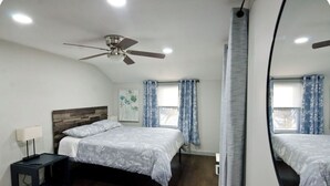 Une chambre à coucher avec un lit, une table de chevet, un ventilateur de plafond et une fenêtre avec des rideaux.