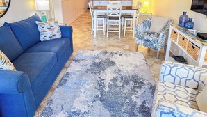 A living room with a blue sofa, a patterned rug, and a dining area with a table and chairs. - Experience Tilghman Beach and Racquet Club Unit 219! 3 Bedroom Ocean View Condo. Pools, Tennis, Close to Main Street! (North Myrtle Beach)