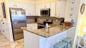 A kitchen with white cabinets, a stainless steel refrigerator, a microwave, a stove, and a granite countertop. There are two blue bar stools and a small table with a lamp. - Experience Tilghman Beach and Racquet Club Unit 219! 3 Bedroom Ocean View Condo. Pools, Tennis, Close to Main Street! (North Myrtle Beach)