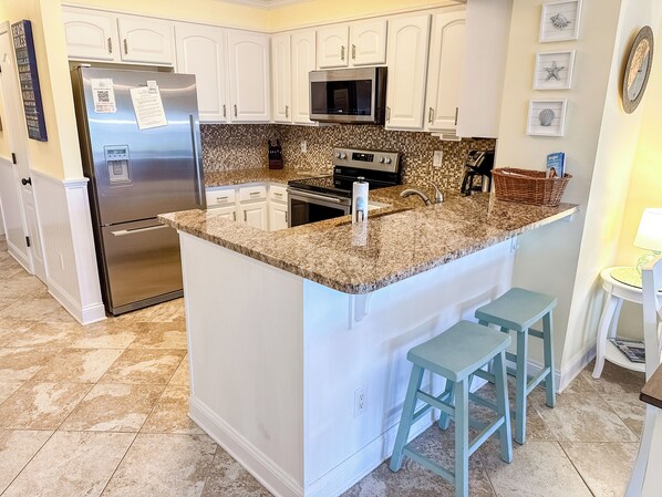 A kitchen with white cabinets, a stainless steel refrigerator, a microwave, a stove, and a granite countertop. There are two blue bar stools and a small table with a lamp. - Experience Tilghman Beach and Racquet Club Unit 219! 3 Bedroom Ocean View Condo. Pools, Tennis, Close to Main Street! (North Myrtle Beach)