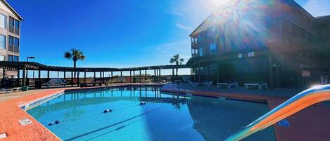 A swimming pool with clear water, surrounded by a red deck area, and a building with balconies in the background.