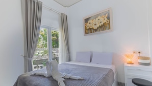 Een net opgemaakt bed met een grijze deken, een wit kussen en een handdoek die bovenop is gevouwen. Aan de muur hangt een schilderij van bloemen, er staat een nachtkastje met een lamp en er is uitzicht op groen buiten.