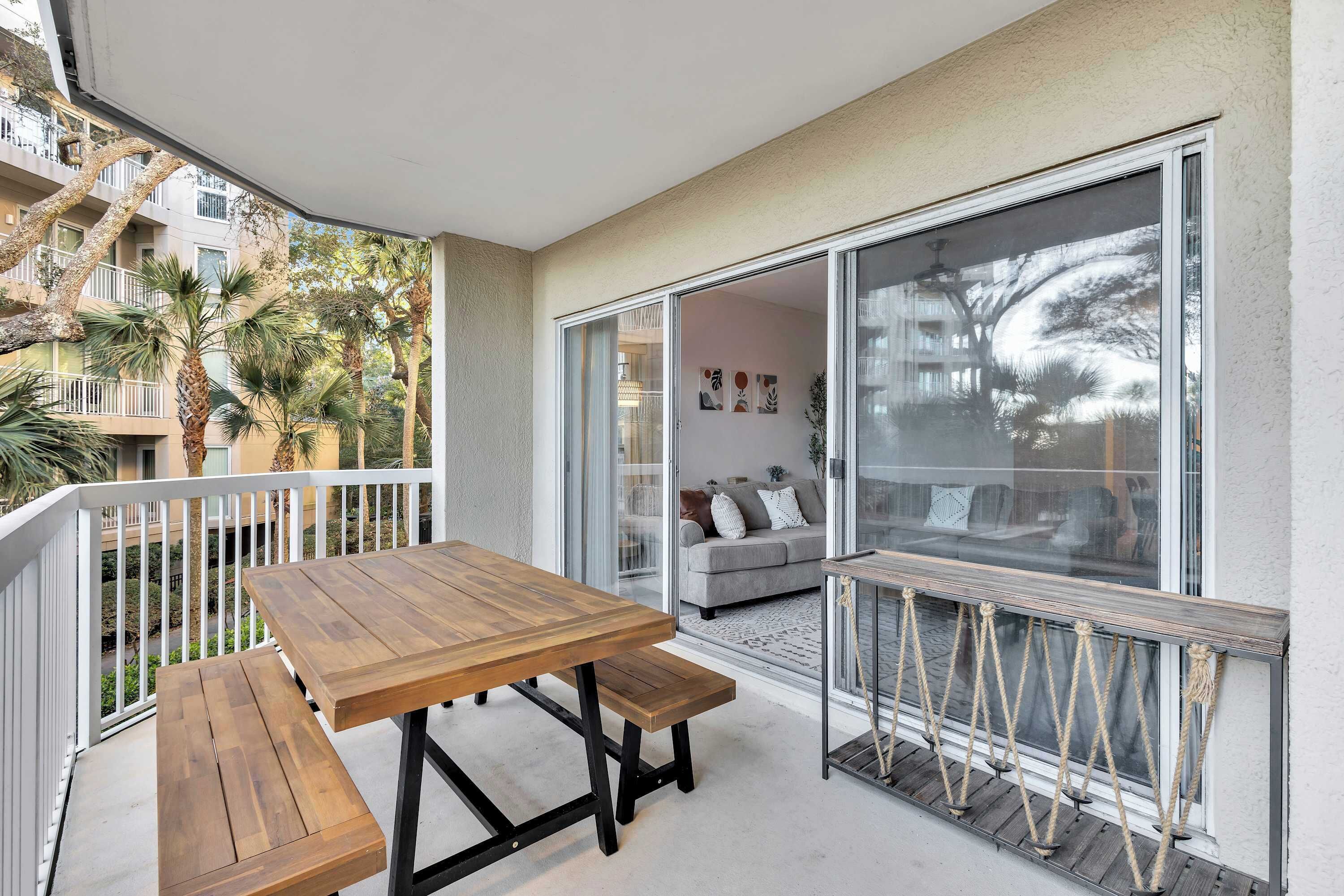 A balcony with a wooden table and bench, a sliding glass door leading to a living area with a sofa and cushions, and a view of palm trees and a building across the street.