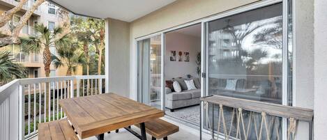 A balcony with a wooden table and bench, a sliding glass door leading to a living area with a sofa and cushions, and a view of palm trees and a building across the street.