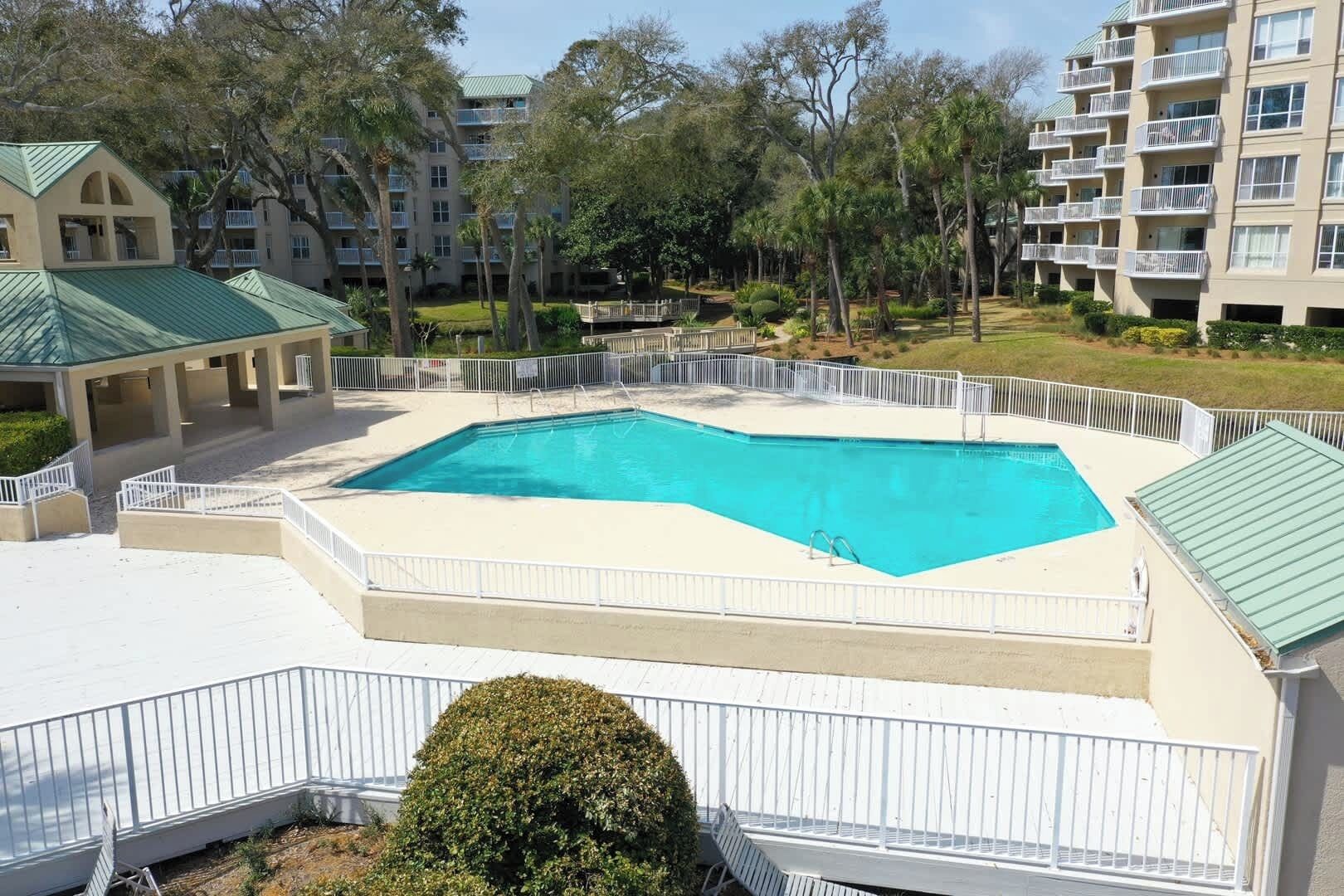 A swimming pool surrounded by a white fence, with a covered area and a building with a green roof in the background.