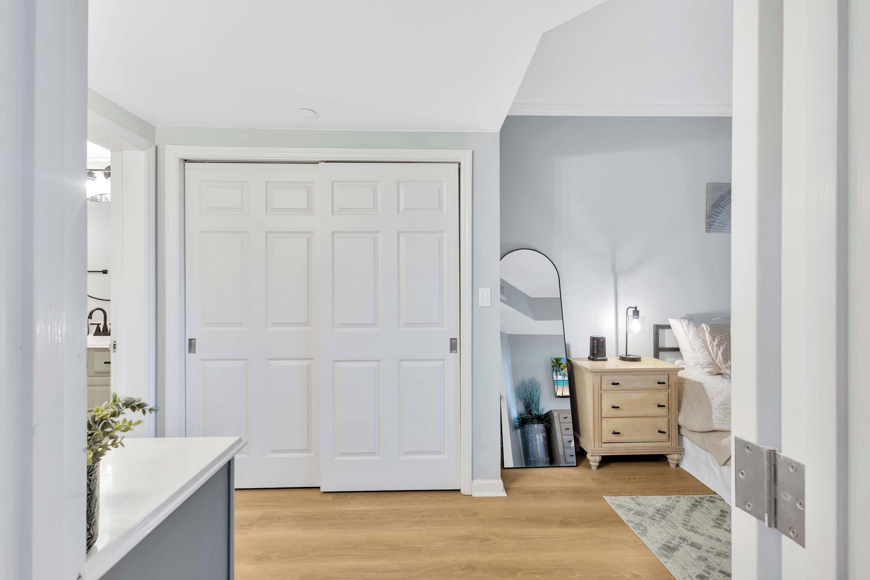 Un couloir avec une porte blanche, une table de chevet avec une lampe, et un lit à l’arrière-plan.