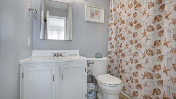 Une salle de bain avec un lavabo, un miroir et des toilettes, tous de couleur blanche. Il y a un rideau de douche orné d’un motif de coquillages.