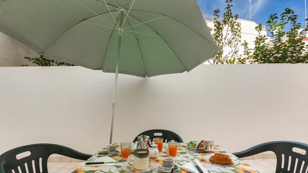 A table set with a floral tablecloth, plates, glasses, and a croissant, under a green umbrella. - 3679 Mono Le Vele (Salve)