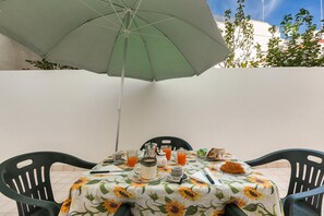 Et bord dækket med en blomstret dug, tallerkener, glas og en croissant, under en grøn parasol.