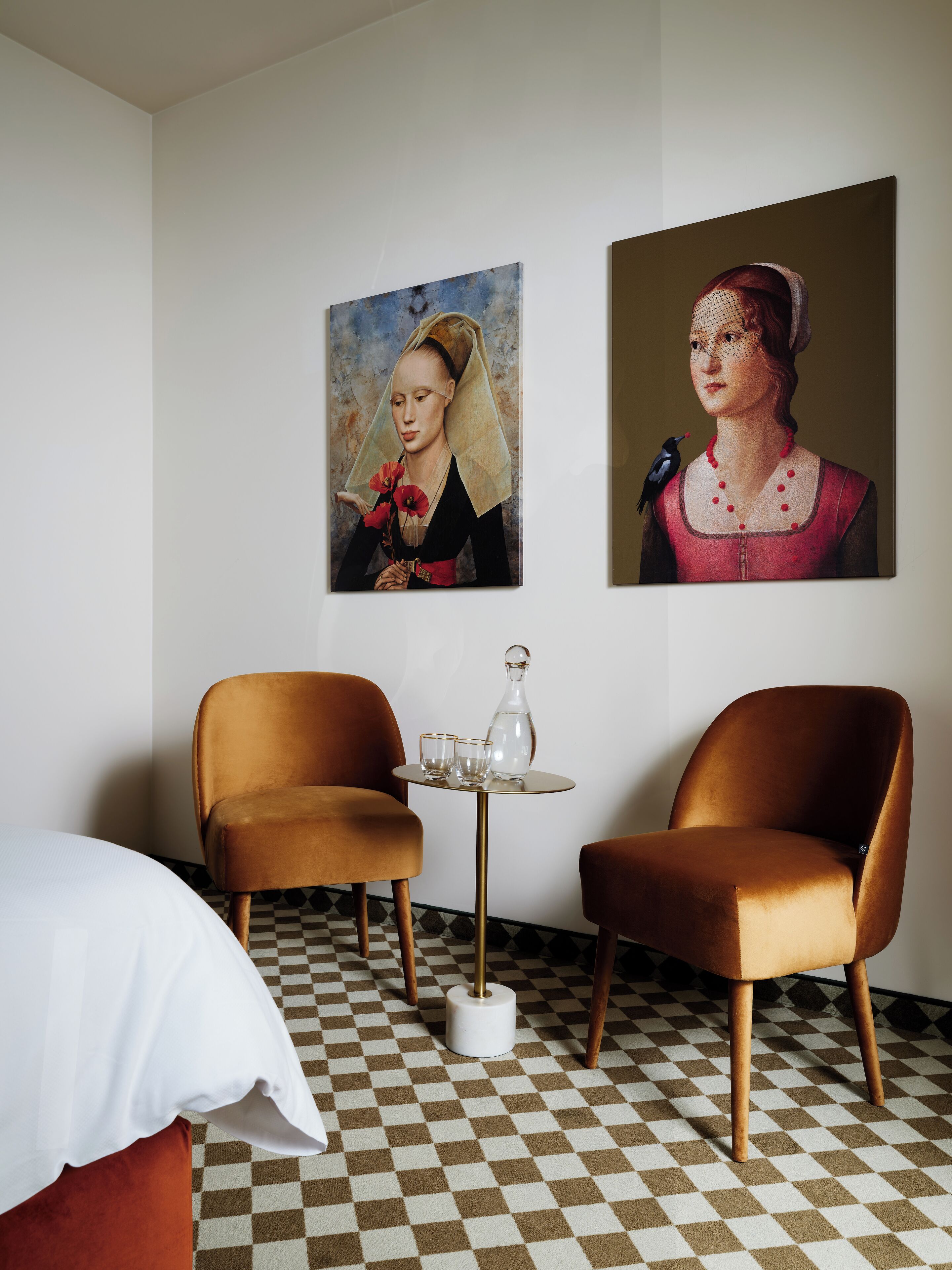 A room with two brown chairs, a small table with a bottle and glasses, and two paintings on the wall.