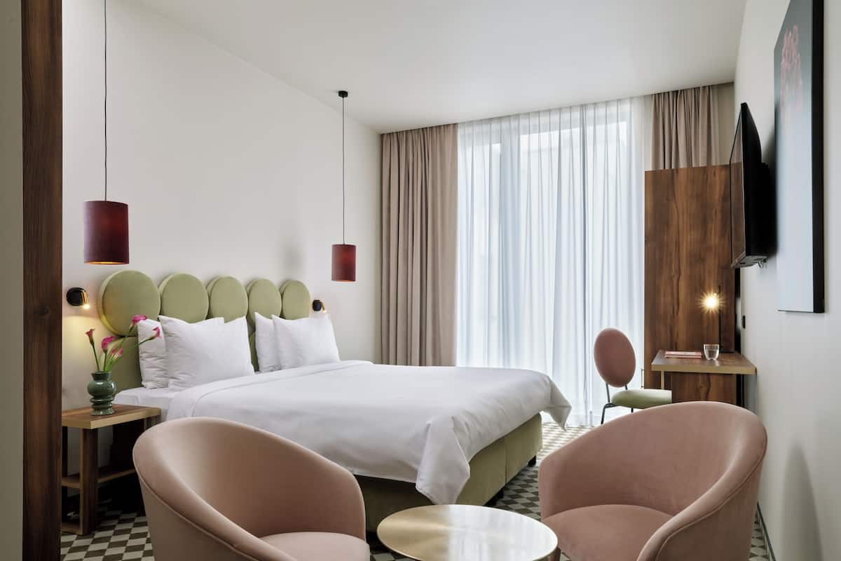 A hotel room with a large bed, two armchairs, a small table, a bedside table with a lamp, a mirror, and a TV mounted on the wall.