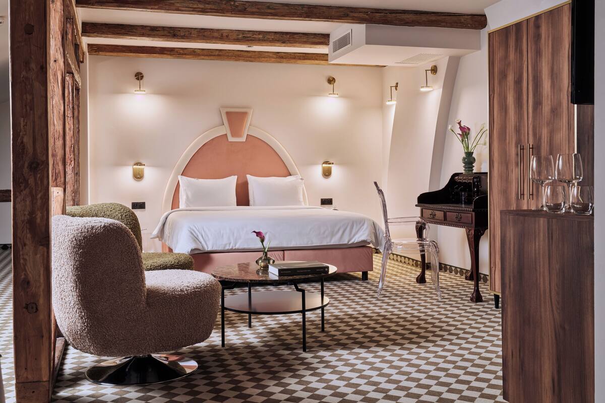 A bedroom with a checkered floor, a bed with white linens, a wooden chair, a small table, and a piano.
