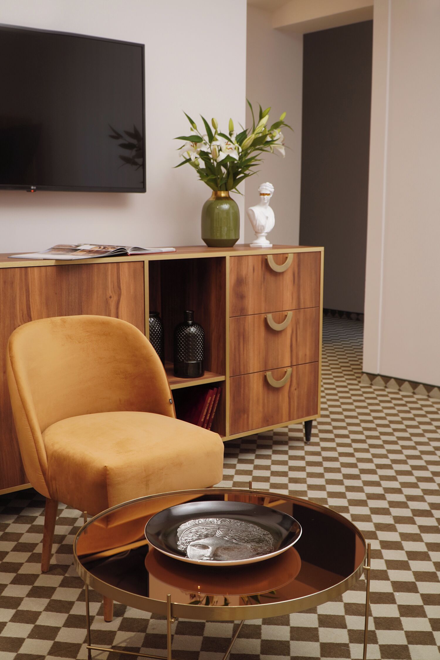 A room with a checkered floor, a wooden cabinet with drawers, a television mounted on the wall, and a round table with a decorative plate.