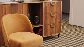 A room with a checkered floor, a wooden cabinet with drawers, a television mounted on the wall, and a round table with a decorative plate.
