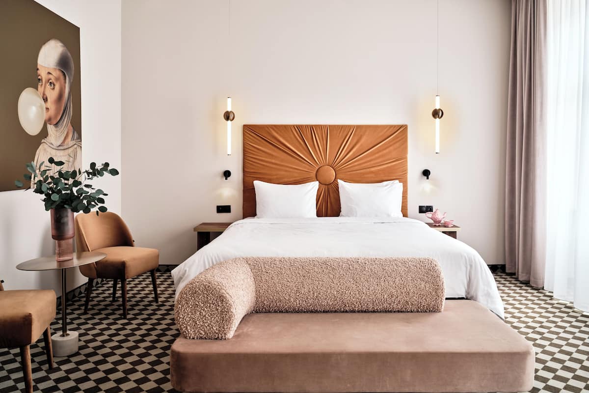 A hotel room with a large bed, a headboard with a circular design, a bedside table with a lamp, a chair, and a potted plant.