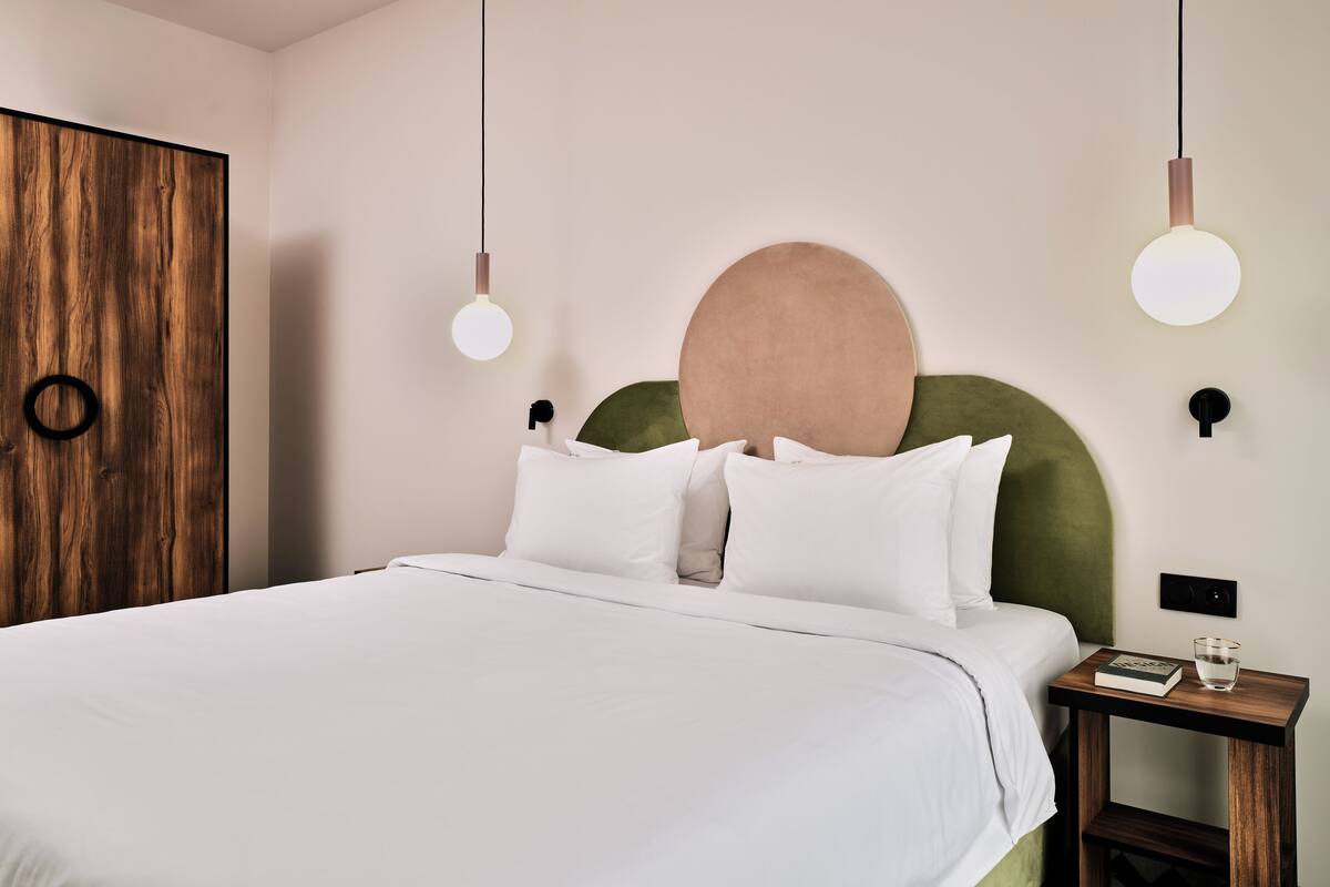 A neatly made bed with white linens, a round headboard, and a bedside table with a book and glass.