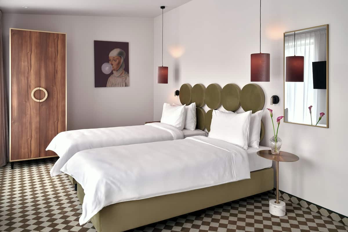 A double bed with white linens, a wooden wardrobe, a painting, and a vase with flowers on a bedside table.