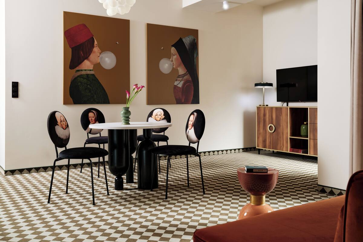 A dining area with a checkered floor, a round table, black chairs, and two paintings on the wall.