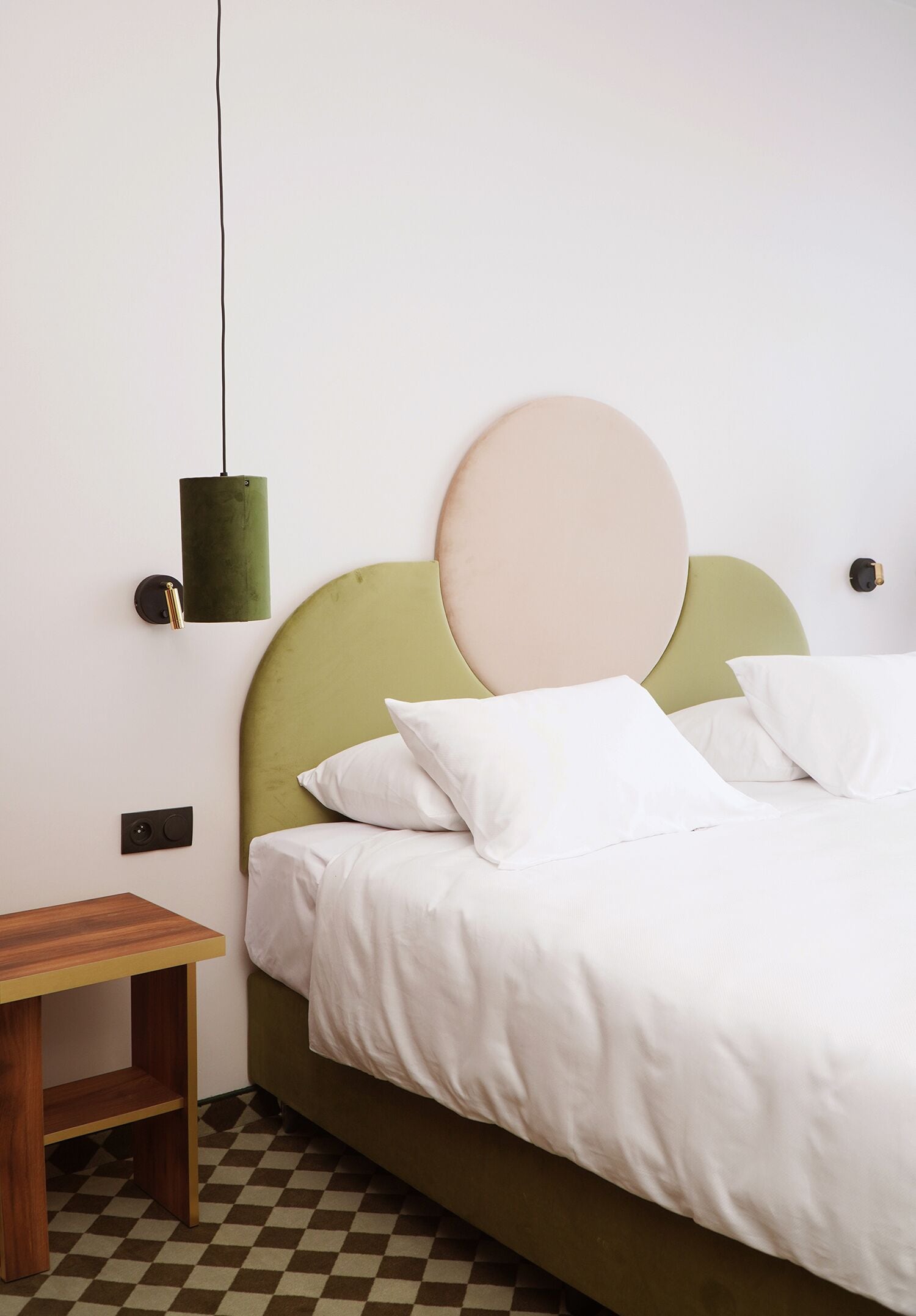 Una cama bien hecha con ropa blanca, un cabecero verde y una lámpara redonda fijada a la pared.