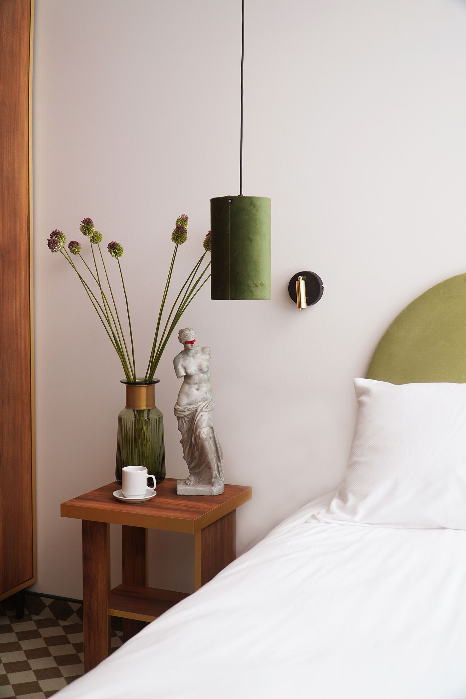 Quarto bem arrumado com uma cama, uma mesinha de cabeceira com um vaso de flores, uma estátua e uma luminária na parede.