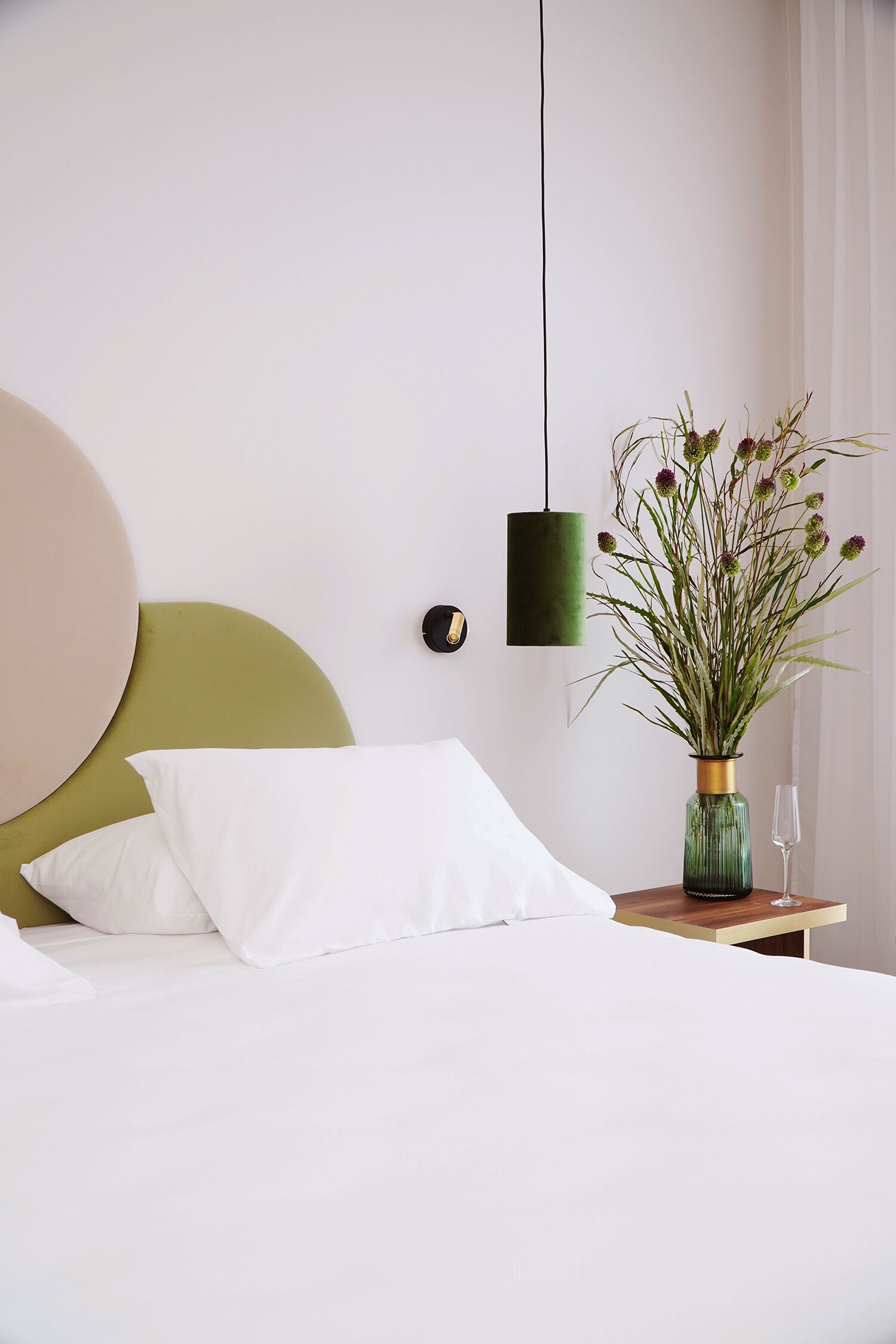 Un lit bien fait, avec du linge de lit blanc, une tête de lit verte et un vase rempli de fleurs posé sur une table de chevet.