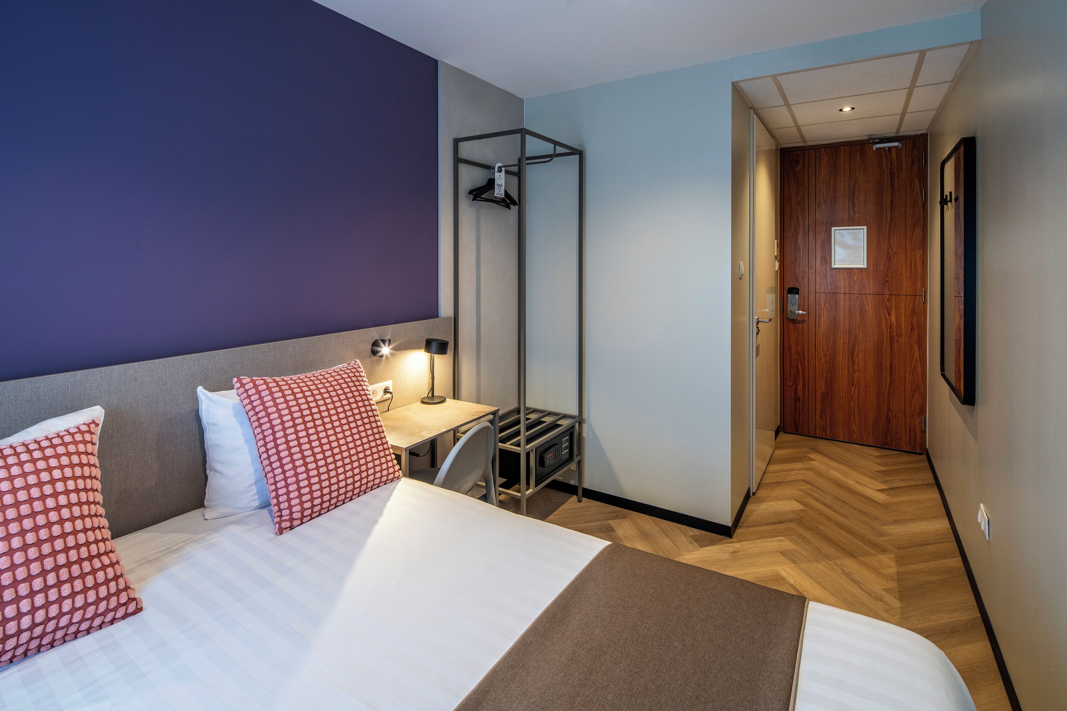Habitación de hotel con cama, almohadas, escritorio con silla, ducha suspendida y puerta de madera.